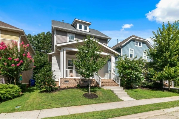 A small tree in front of a house with a brick porch. It is sunny outside and there is a sidewalk in front of the house.