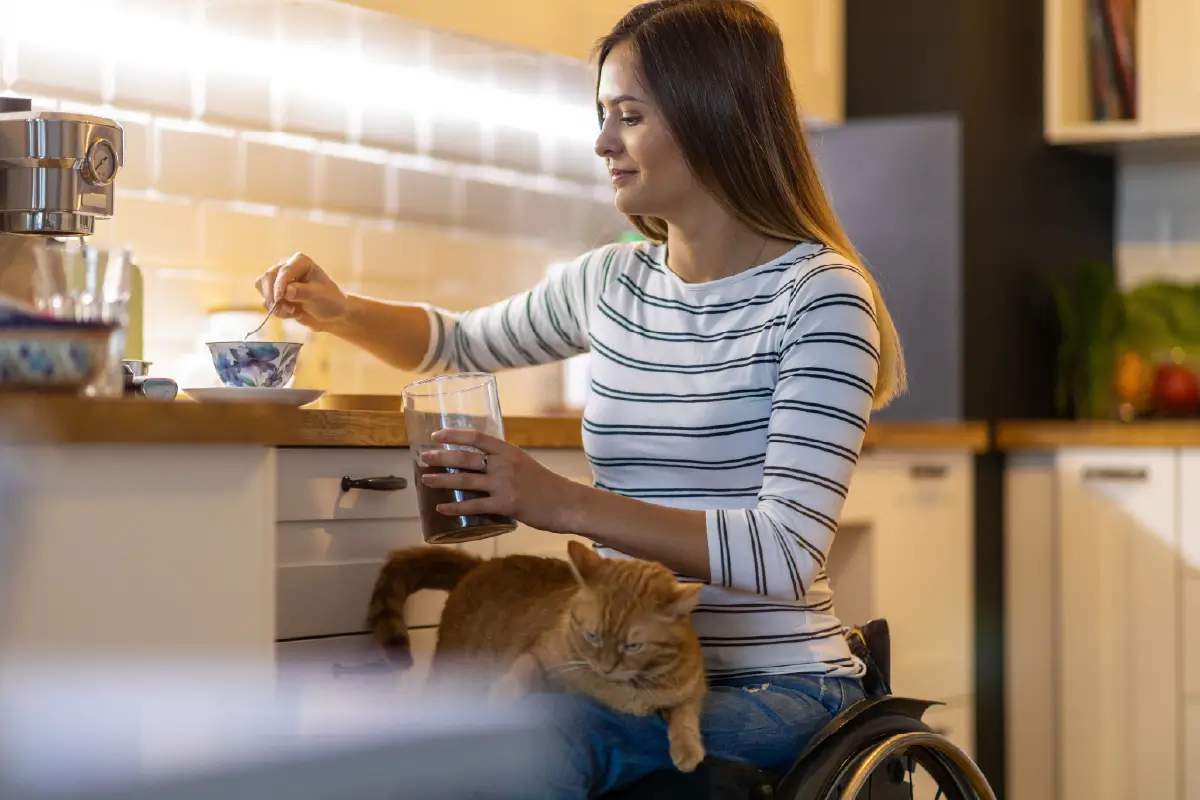 Photograph of a woman using a wheelchair in the kitchen, preparing a cup of tea. An orange cat is on her lap.