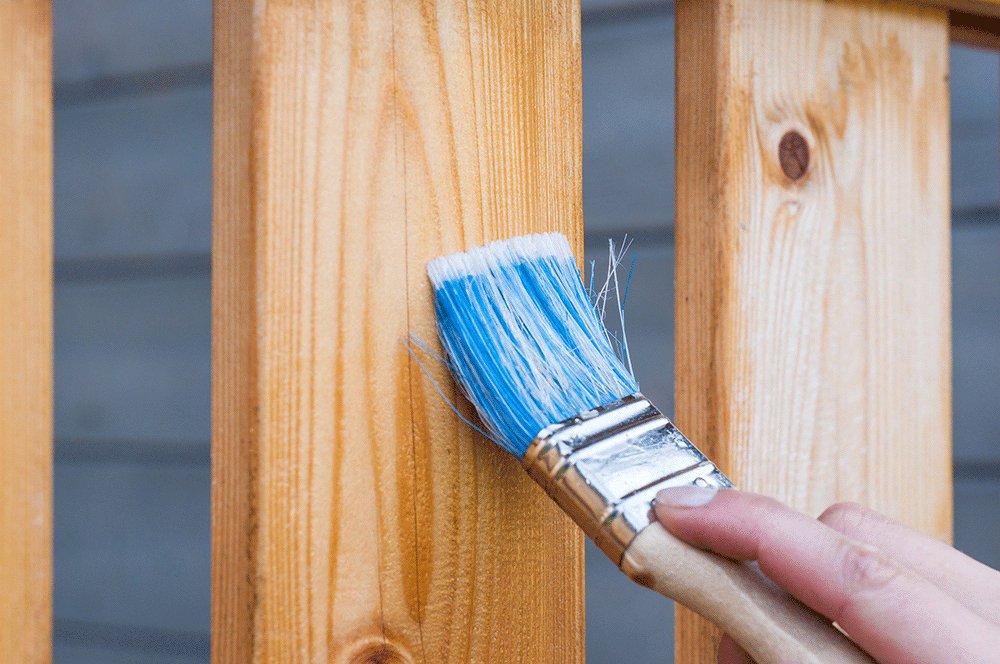 Hand holding a paintbrush and applying stain to a piece of wood on a deck railing.