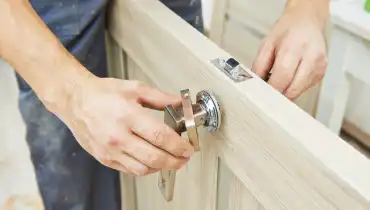A handyman is holding a door slab between his legs as he adjusts the door handle while providing door repair services.