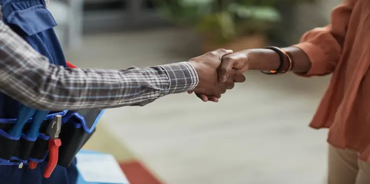 Person wearing bracelets and a salmon-colored shirt shaking hands with person wearing plaid flannel shirt and toolbelt.