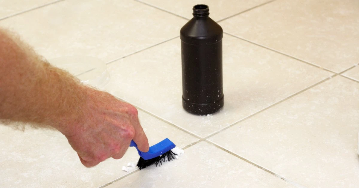A handyman using a grout brush and grout cleaner to clean the grout lines on bathroom floor tiles while providing services for bathroom maintenance.