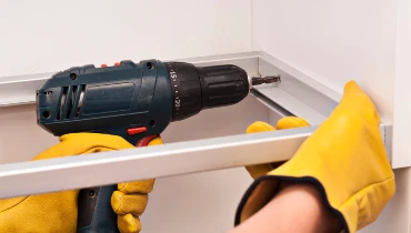 A close-up image of a handyman using a power drill to mount cabinet boxes on a wall during a cabinet installation project.