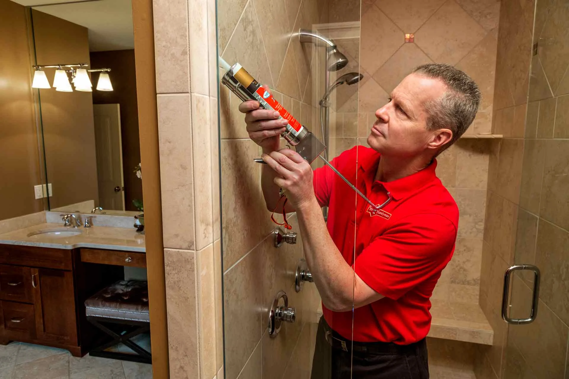 A handyman using a caulking gun to add fresh caulk to the joint between a glass shower door and a tile shower wall.