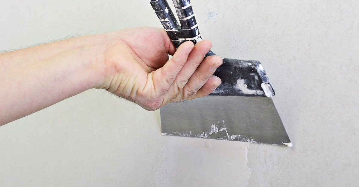 A handyman using a wide putty knife to spread joint compound over a hole in a wall during drywall repairs.