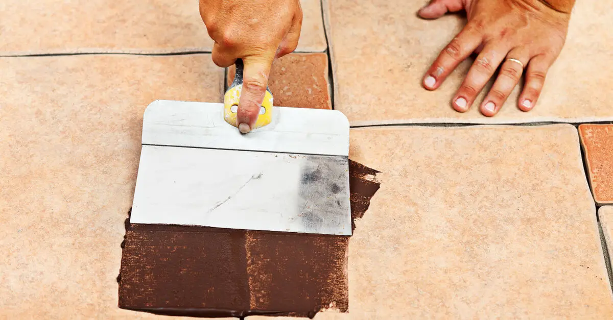 A handyman using a grout float to apply brown grout to the gaps between beige tiles on a floor.