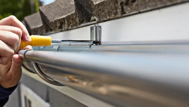 A handyman using a screwdriver to complete gutter installation along a residential roof.