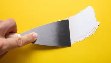 A close-up of a putty knife being used to spread spackle across a yellow wall that is receiving drywall repairs.