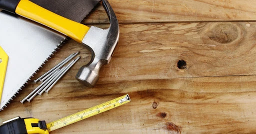 A hammer, saw, tape measurer, and nails laying on top of wooden planks before they’re used for floor repair.