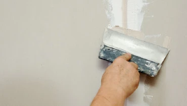 A handyman using a putty knife to spread joint compound over a taped seam between two pieces of drywall.