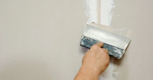 A handyman using a putty knife to spread joint compound over a taped seam between two pieces of drywall.
