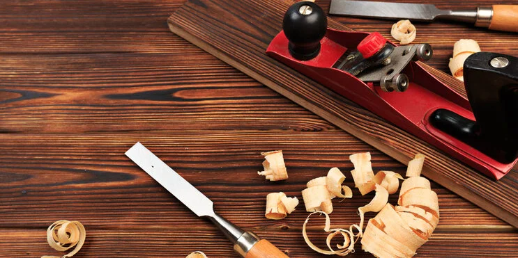 A closeup of two chisels and a wood plane used for interior carpentry work, surrounded by ribbons of wood and on top of a wood surface.