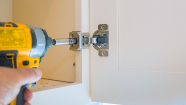 The bottom corner of a cabinet where a handyman is using a drill to adjust the door hinge while providing cabinet repair services.