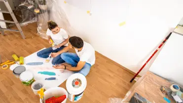 A man and a woman sit on the floor while testing multiple colors of paint for their home using a sheet of paper.