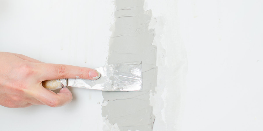 A close-up of a hand holding a small putty knife as it is used to apply joint compound to a wall during a drywall repair project.