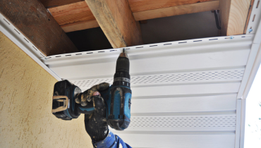 A handyman using a power drill to provide soffit repair services along a residential roofline.