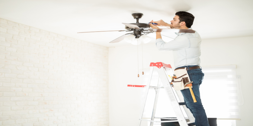 A handyman with electrical skills and a tool-belt stands on a ladder while fixing a ceiling fan