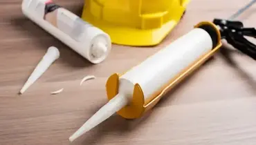 A tube of caulk loaded in a caulking gun and another empty, used tube laying next to a hardhat nearby