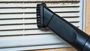 A close-up of a vacuum cleaner nozzle sucking dust and lint off a ventilation cover.