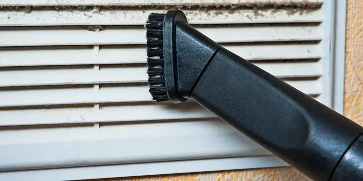A close-up of a vacuum cleaner nozzle sucking dust and lint off a ventilation cover.