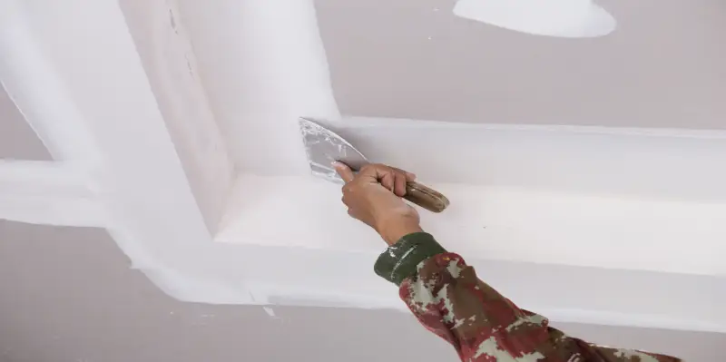 Person's hand using a putty scraper on a ceiling that has recently had its drywall repaired.