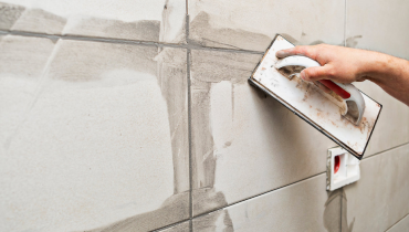 A handyman using a grout float to apply gray grout to the joints between white tiles in a tile backsplash.