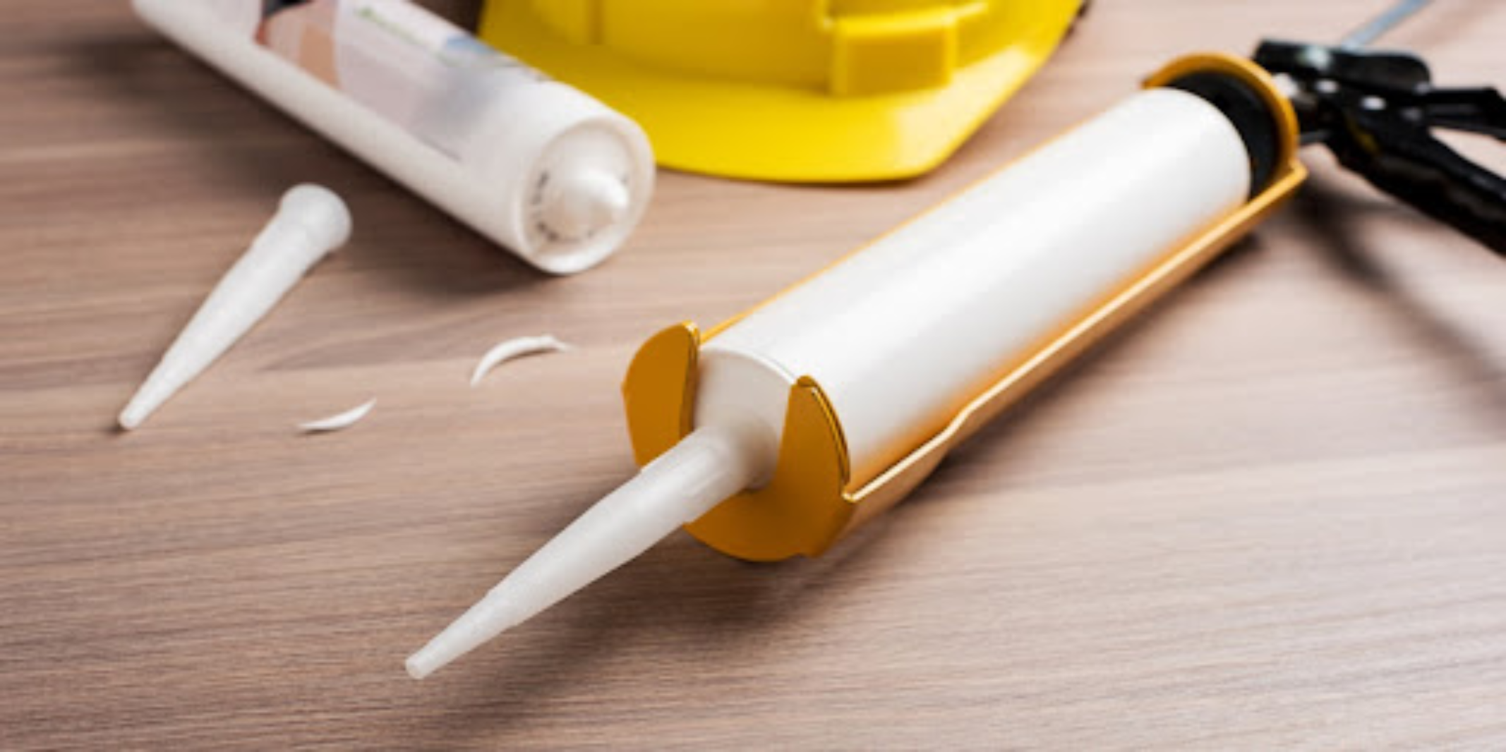 A caulking gun with a full tube of fresh caulk lying on a table next to an empty tube and a yellow hard hat.