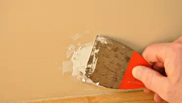 A handyman using a putty knife to spread spackle over a small hole that has been patched with drywall repairs.