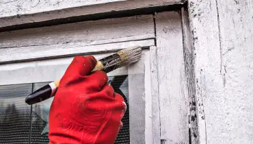 A gloved hand holding a paintbrush as a handyman repaints the outer trim around a window after completing trim repairs.