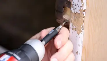 A handyman using a power drill to repair the strike plate for the latching mechanism of a residential door.