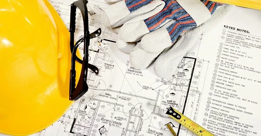 A hardhat, safety glasses, work gloves and the end of a tape measurer lying on top of detailed home remodeling plans.