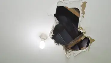 A hole in a residential ceiling located near a light fixture, with wooden ceiling beams visible in the space beyond the hole