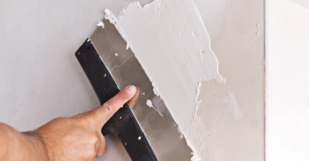 A handyman using a wide putty knife to spread drywall compound across a wall while completing drywall repairs.