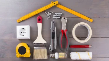 Tools like a tape measure, pocketknife, paintbrush, and nails on a wood surface arranged in the shape of a house.