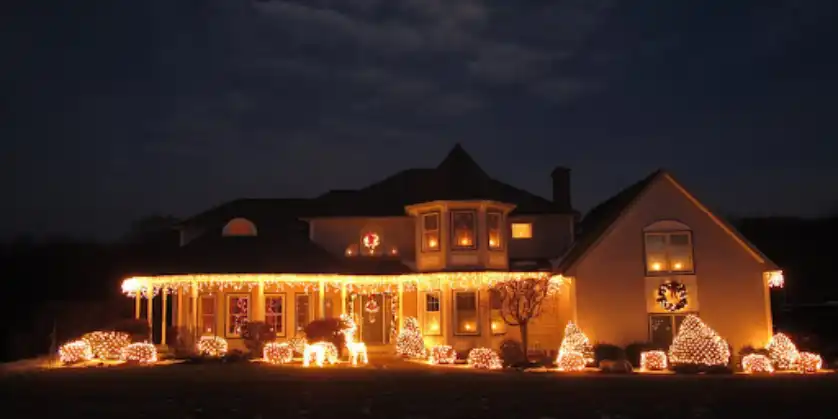 A two-story home with bright holiday lighting hanging from the lower roofline and awning over the front deck, as well as wrapped around foliage in the front yard.
