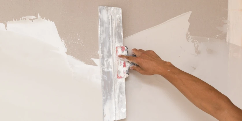 A close-up of a handyman using a large putty knife to spread joint compound over a wall where they have recently completed drywall repairs.