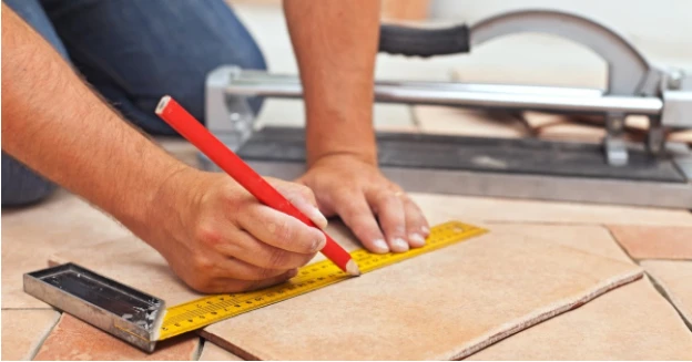 Person's hands using measuring tape and pencil to mark a line on a loose piece of tile on the ground.