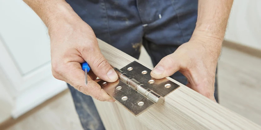 A handyman lining up a new hinge on the side of a door slab during an appointment for door repairs.