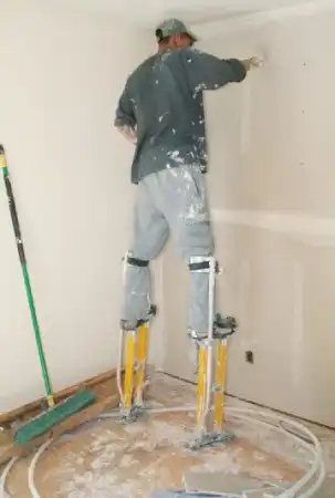 Man standing on yellow stilts to repair drywall near the top of an interior wall.
