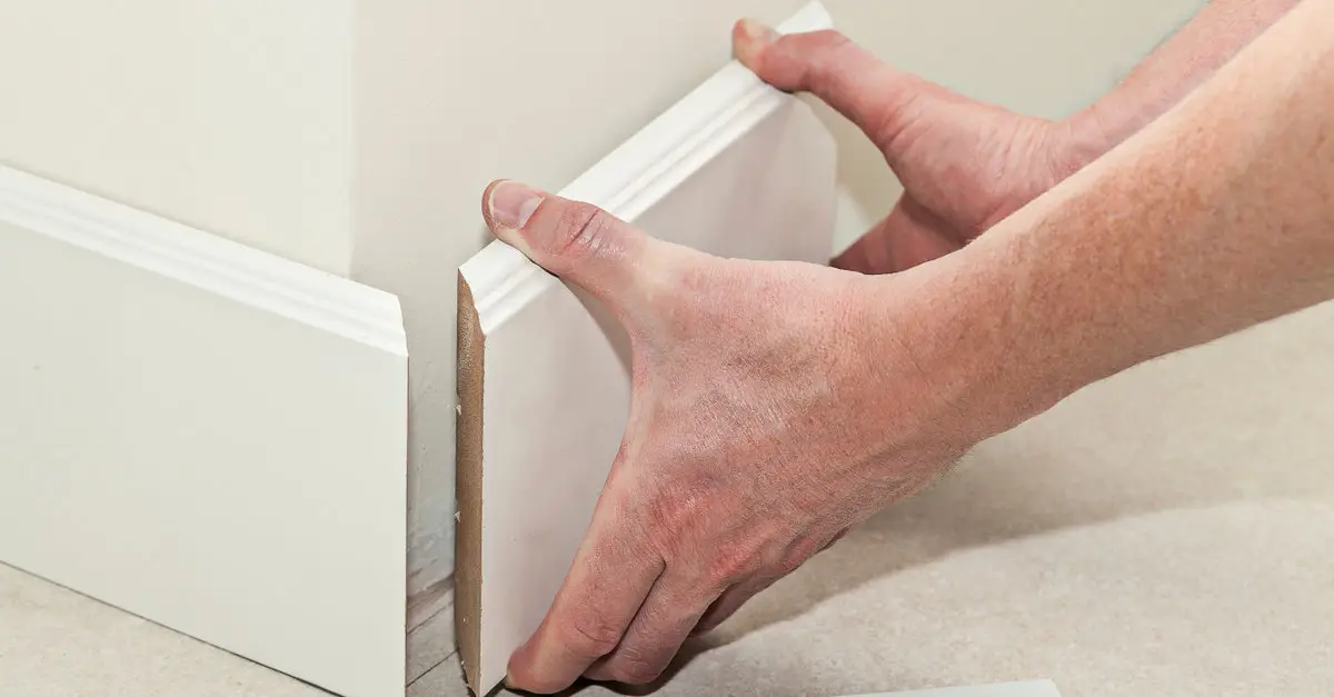 A close up of a handyman’s hands as he installs a new section of baseboard trim along the base of a wall.