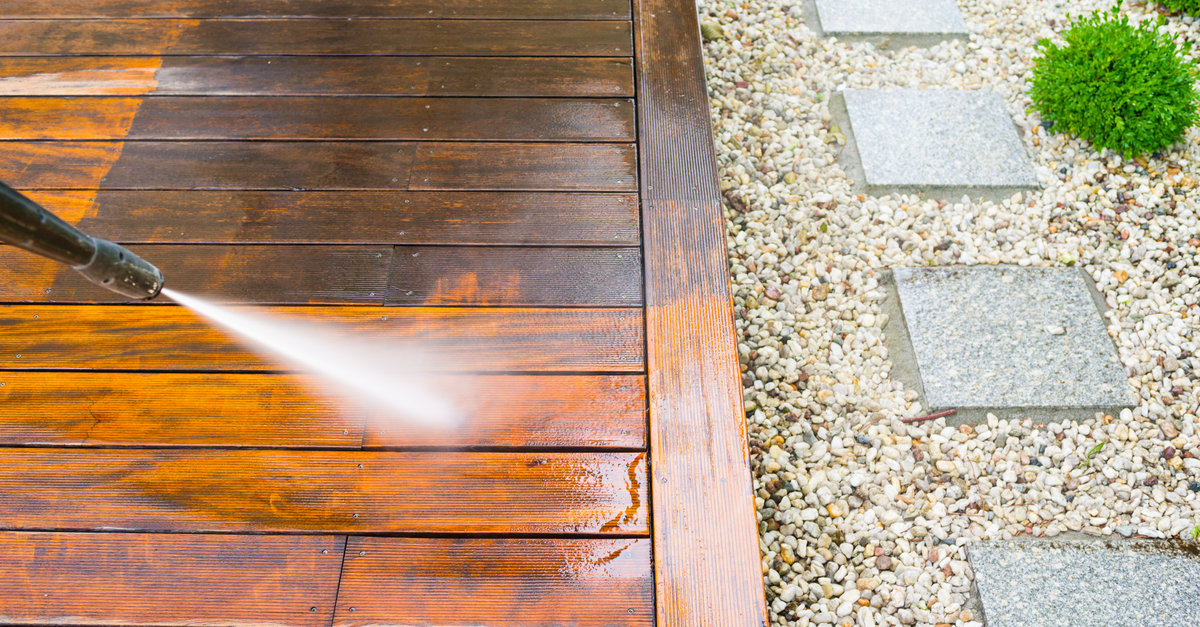 A pressure washer blasting a jet of water at a wooden deck while a handyman provides pressure washing service to clean away a layer of grime on the deck.