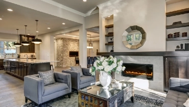 Chic living room filled with built-in cabinets flanking round mirror atop grey tile fireplace, tufted sofa facing two armchairs and window wall overlooking lush outdoors