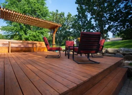 The sun shines on a deck with a fresh coat of stain, a pergola, and chairs.