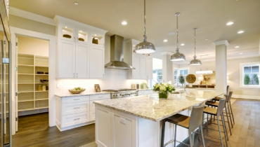 White kitchen design features a large bar style kitchen island with granite countertop illuminated by modern pendant lights. Open doors lead to a walk-in pantry.