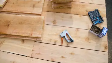 A handheld, manual nail gun, a box of nails, drill bits, and new wooden boards all lying on a wooden porch in preparation for porch repairs in Wichita, KS.