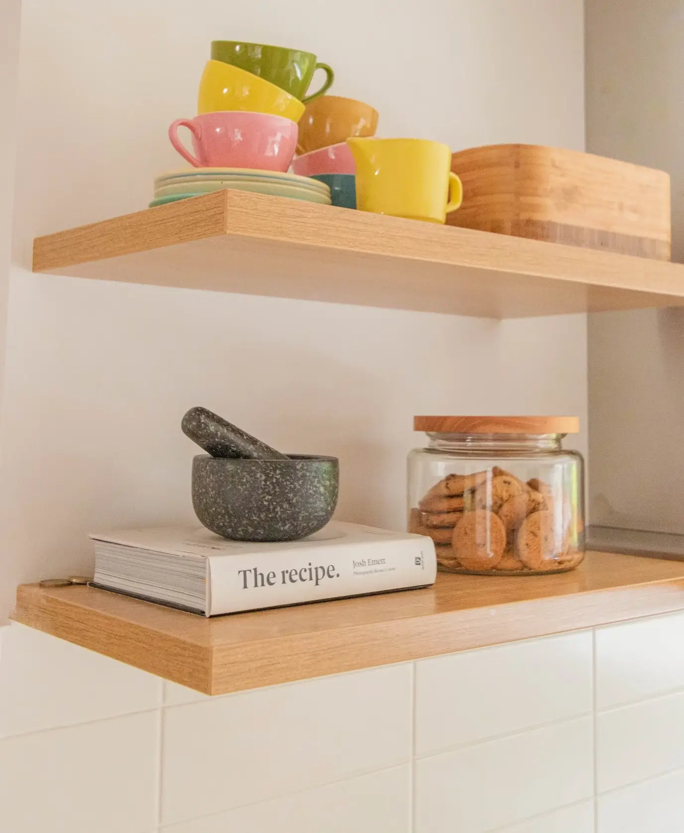 Two wooden shelves are on a white wall. The top shelf has colorful cups and saucers set on it, as well as a wooden box. The lower shelf has a recipe book, a mortar and pestle, and a jar of cookies on it.