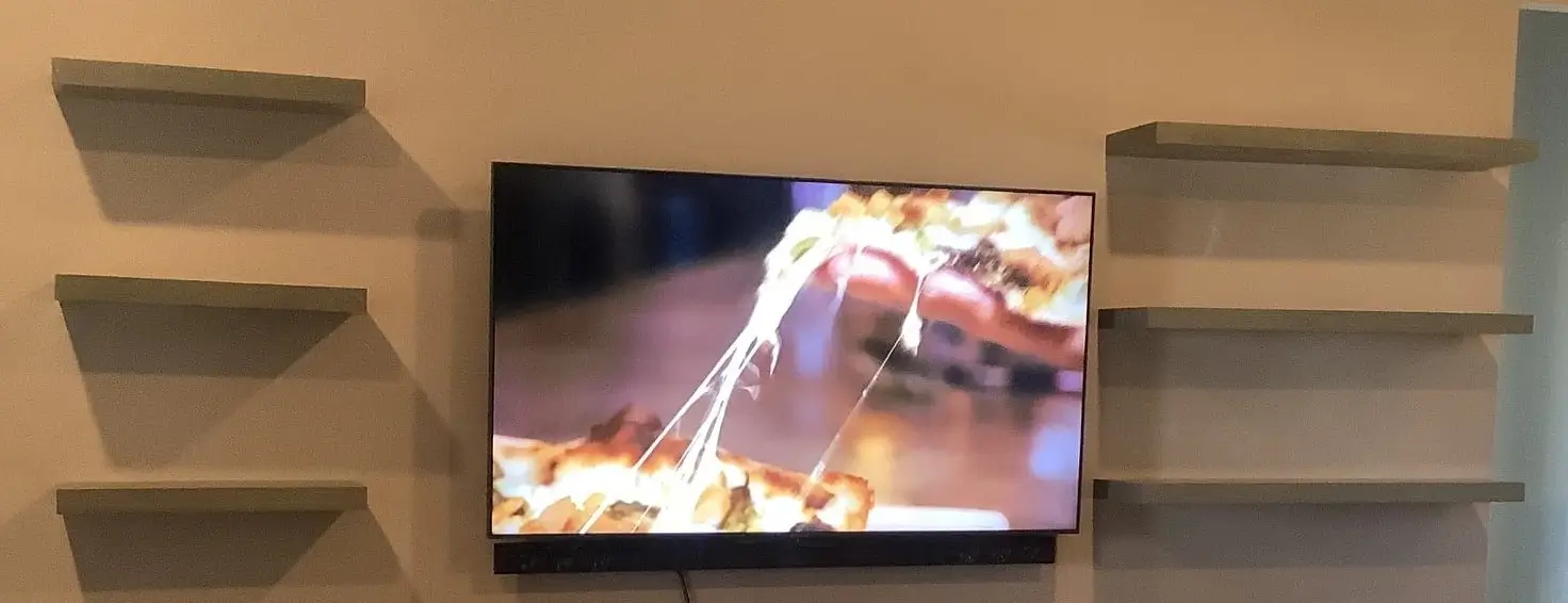 A tan wall holds 6 newly installed shelves, 3 on either side, of a flat screen tv that is hanging in the middle. The tv has an image of a cheesy piece of pizza on it.