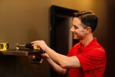 A Mr. Handyman service professional in a red shirt drills a shelf into a wall with a level tool set on top of the shelf.