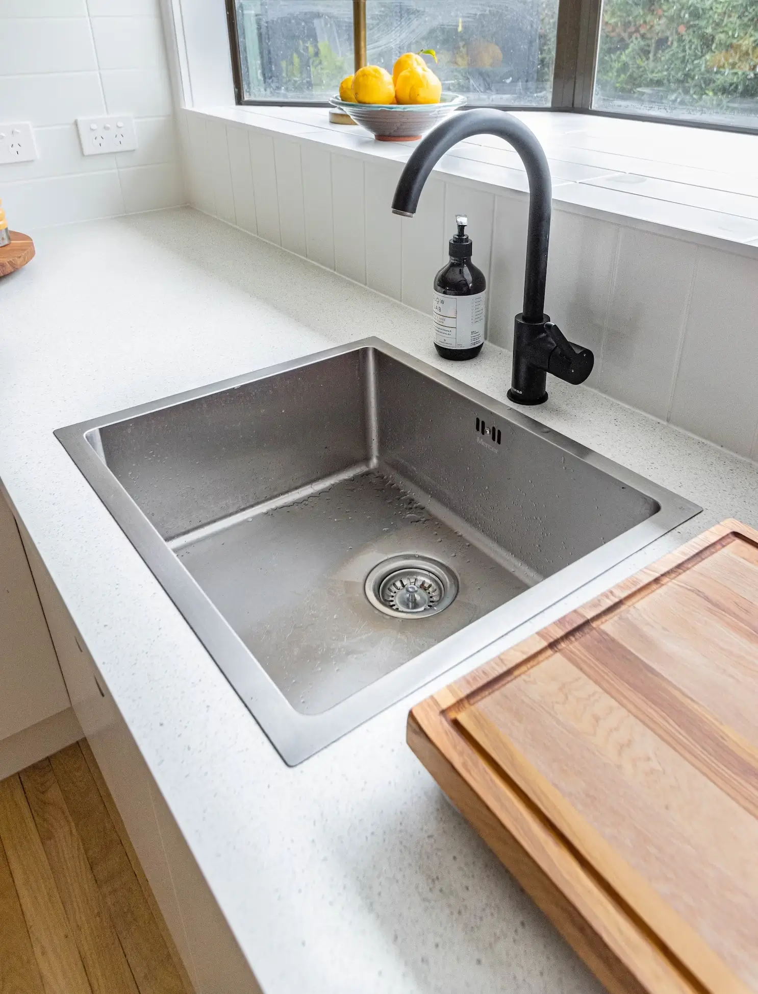A stainless steel sink installed in a white countertop. There is a bowl of lemons set behind the sink and a window that looks out from the kitchen.