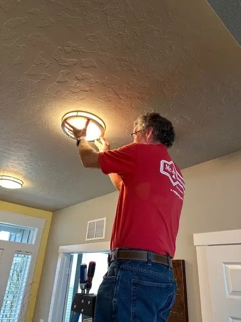 A man wearing a red Mr.Handyman shirt is installing a round light fixture into the ceiling.
